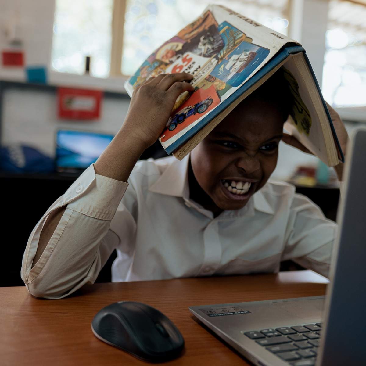 A frustrated student holding a book on their head while looking at a laptop